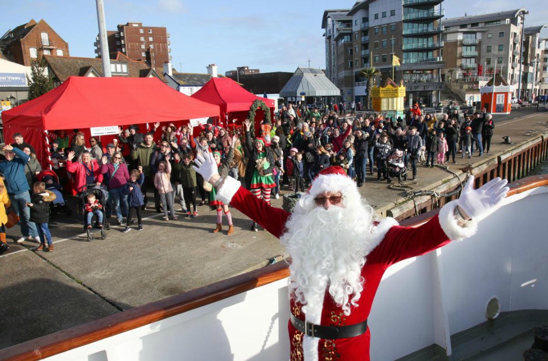 Santa standing on a boat on a sunny day at Poole Quay with a large crowd of families in the backgroudn